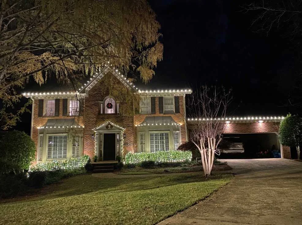 Home with warm white roofline Christmas lights, lit landscaping, and a decorated tree at night