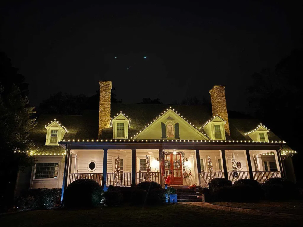 Large traditional home with warm white roofline Christmas lights, wreaths, and tall chimneys