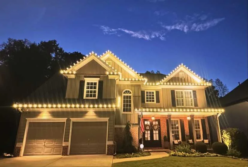 Large home with warm white Christmas lights along the roofline and front entry at dusk