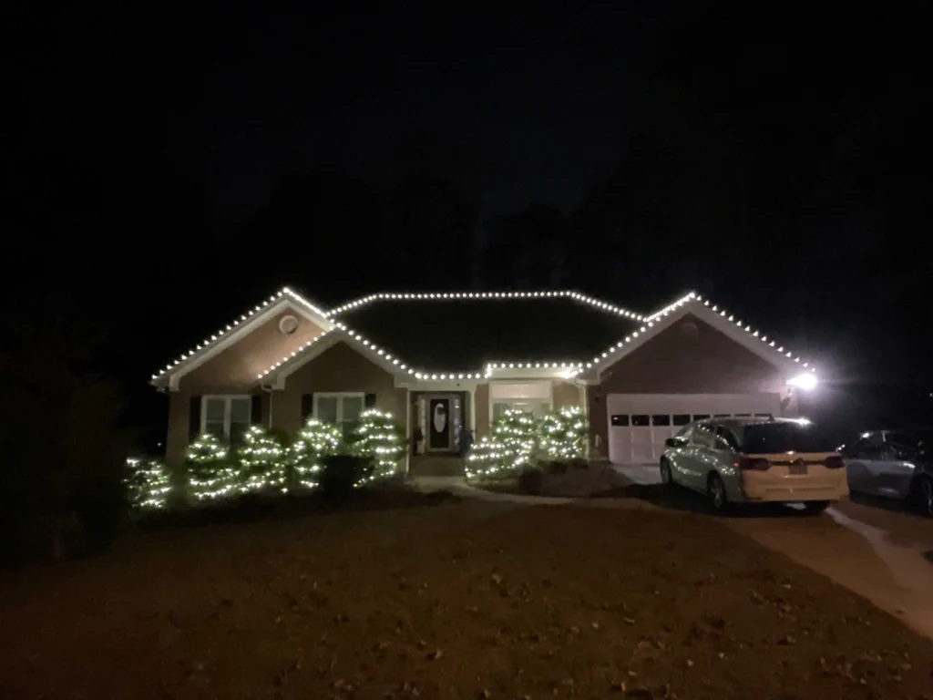 Single-story home with warm white roofline lights and illuminated shrubs across the front yard