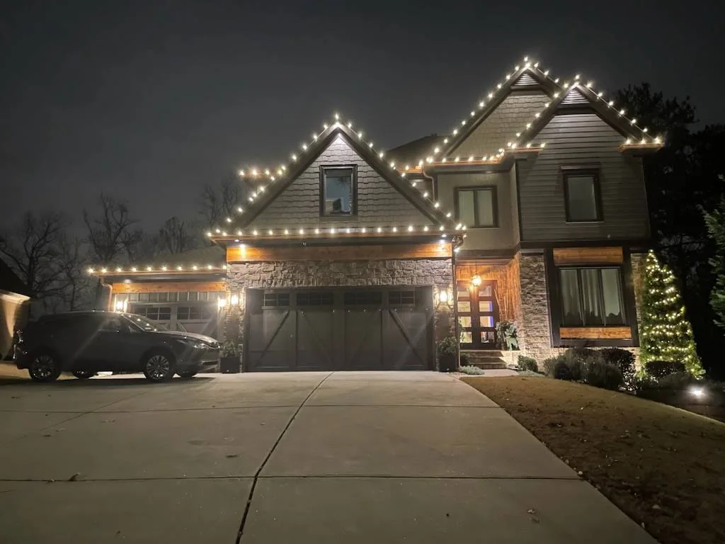 Two-story home with warm white Christmas lights outlining roof peaks and the garage